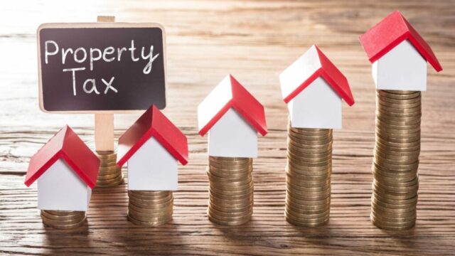 Coin stacks with small house models on top and a chalkboard sign reading "Property Tax".