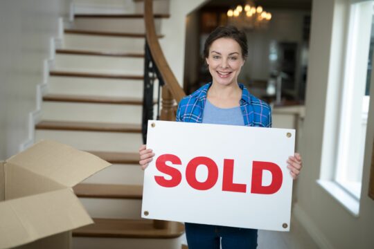 Sold sign with happy woman in new home holding box and moving-in process, real estate transaction, house sale, home ownership, property sale, mortgage, and lending services, Mortgage Factory™.